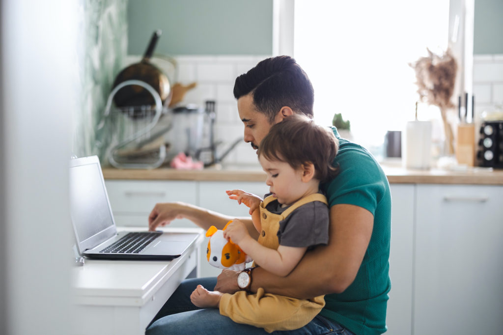 Photo of a dad holding infant sitting at laptop