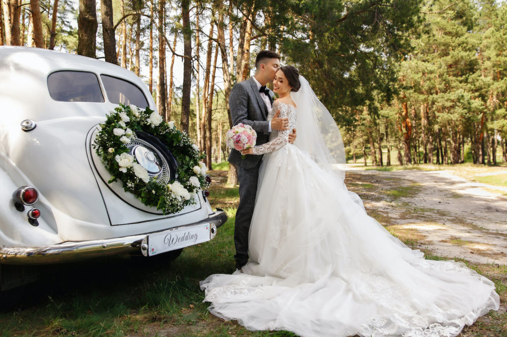 Luxury wedding photography. Elegant wedding couple, groom in grey suit and bride in wedding dress with long sleeves and long train kissing near white Just Married car with inscription "Wedding"