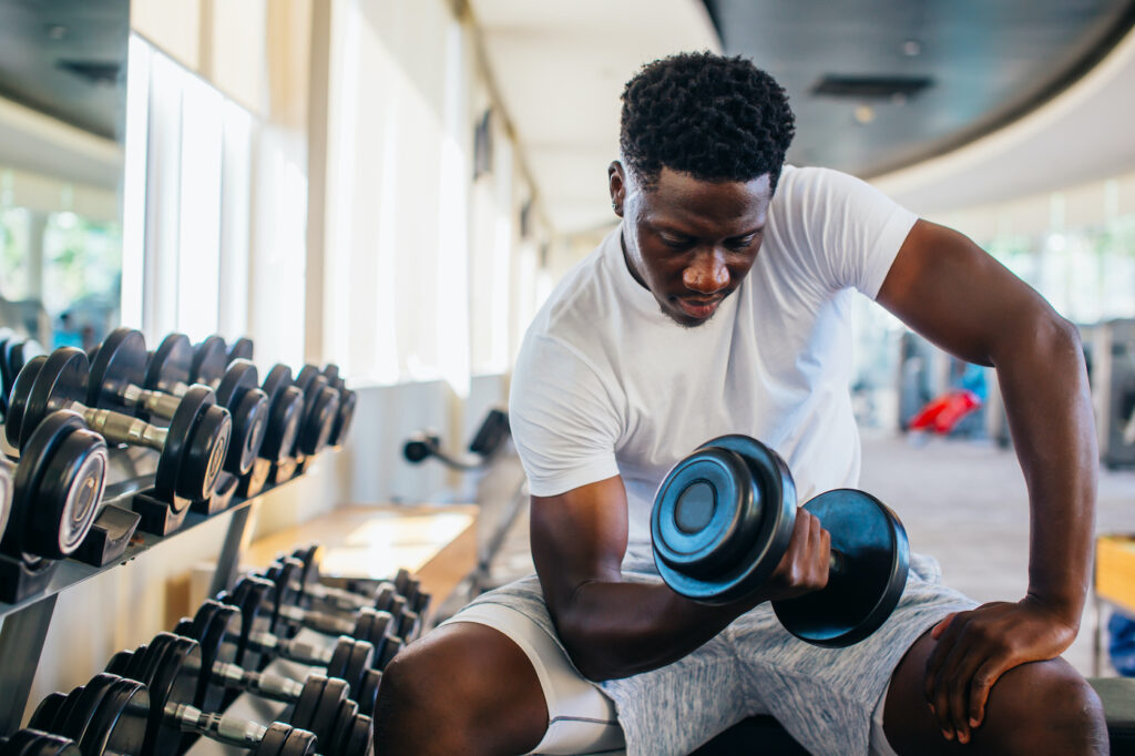 Young African American man sitting and lifting a dumbbell with the rack at gym
