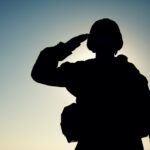Silhouette of soldier in combat helmet and ammunition saluting on background of sunset sky. Army special forces fighter
