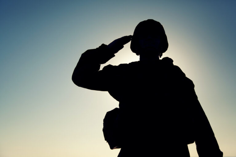 Silhouette of soldier in combat helmet and ammunition saluting on background of sunset sky. Army special forces fighter