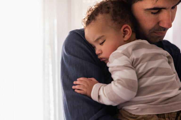 Father hugging son with eyes closed besides a window while spending time at home