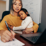 Young working mom writing notes while embracing her daughter on her lap. Multi-tasking mom making business plans while sitting at her desk. Mother of one working remotely in her home office.