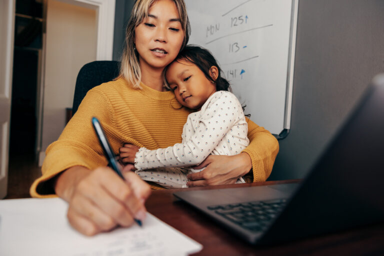 Young working mom writing notes while embracing her daughter on her lap. Multi-tasking mom making business plans while sitting at her desk. Mother of one working remotely in her home office.